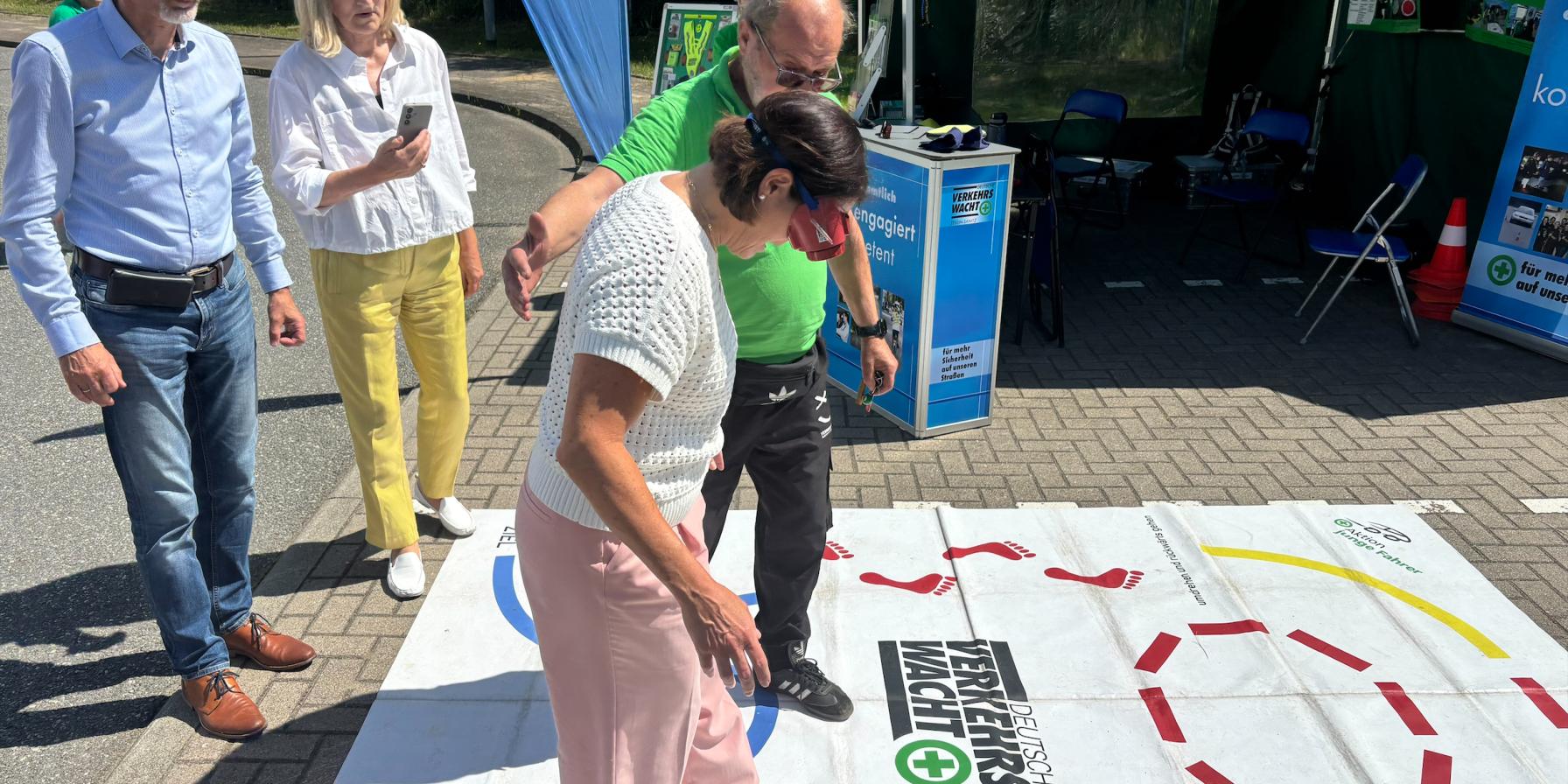 A woman wearing vision-impairment goggles navigates a road-safety activity mat with guidance from an instructor, while two people watch nearby at an outdoor safety event.