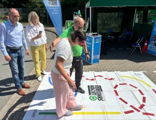 A woman wearing vision-impairment goggles navigates a road-safety activity mat with guidance from an instructor, while two people watch nearby at an outdoor safety event.